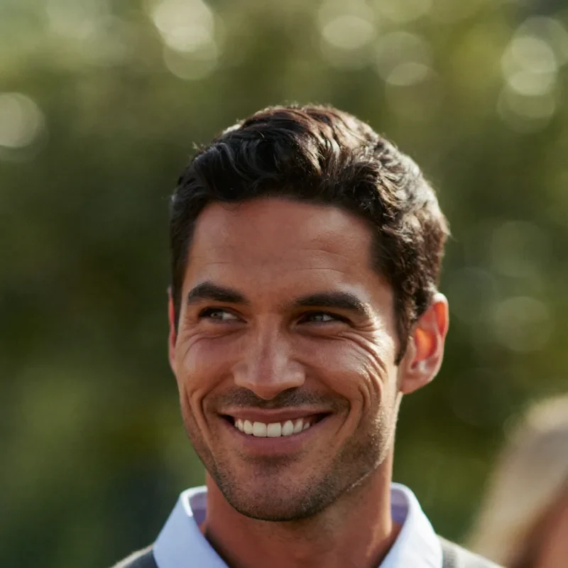 Ritratto di un uomo sorridente con maglione grigio e camicia azzurra, su sfondo naturale sfocato